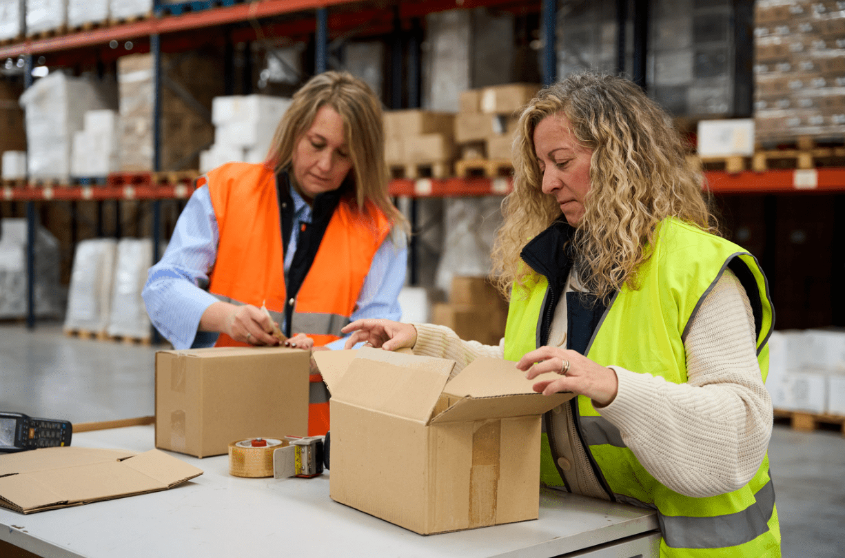 Two women in a warehouse with warehouse packaging supplies including boxes, and tape.